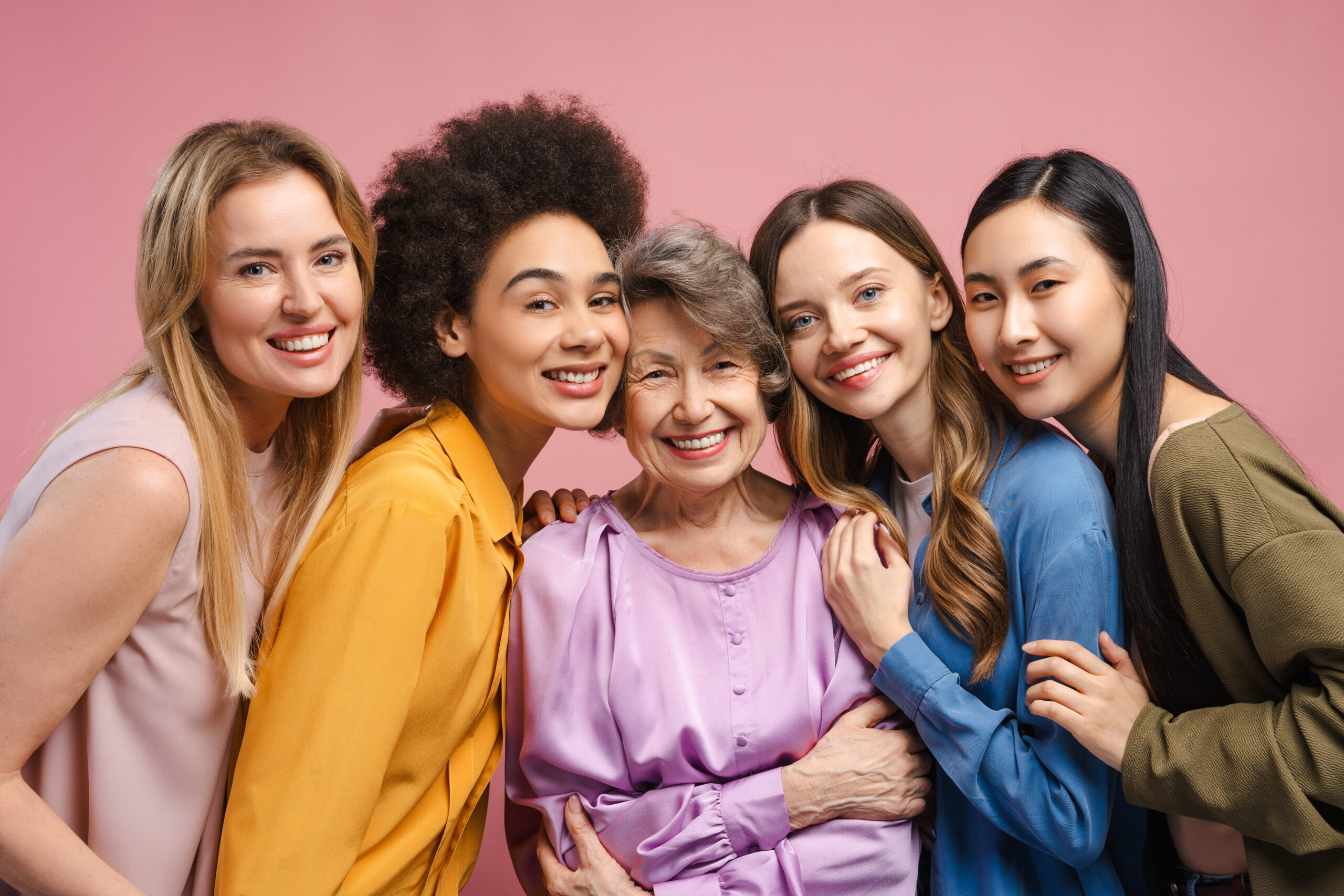 Five cheerful multi-ethnic women of varying ages hugging an elderly woman against a pink background, celebrating their diverse friendship with smiles and joy